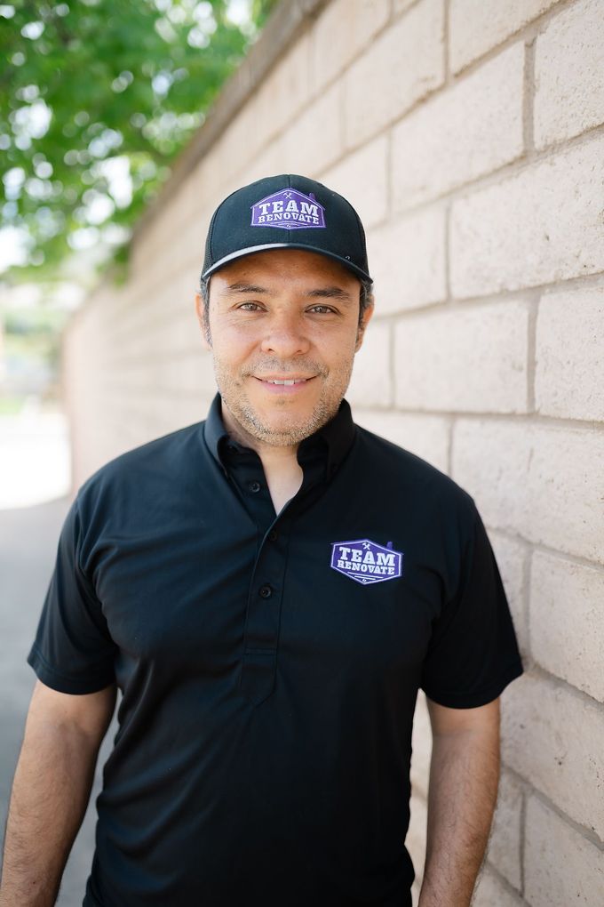 A man wearing a hat and a black shirt is standing in front of a brick wall. Owner at Team Renovate in San Diego , Home remodeler