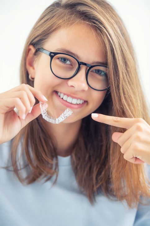 A young woman wearing glasses is holding a clear brace on her teeth.