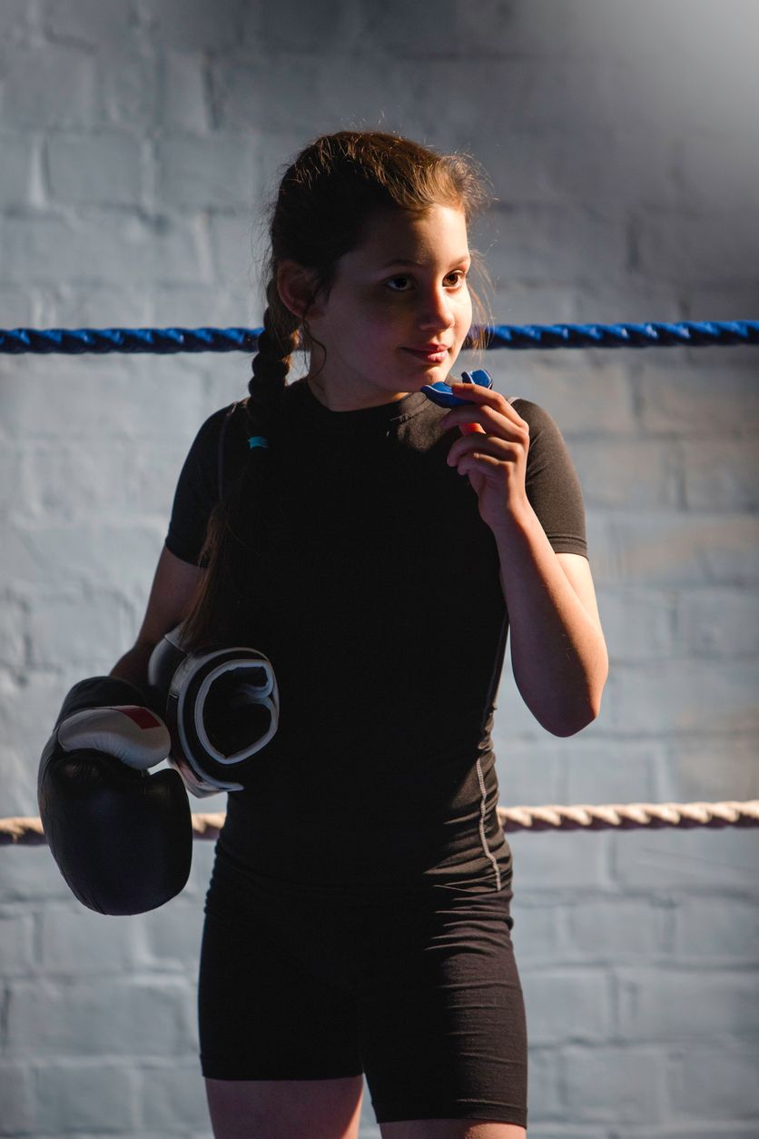 A young girl is standing in a boxing ring holding a mouth guard and boxing gloves.