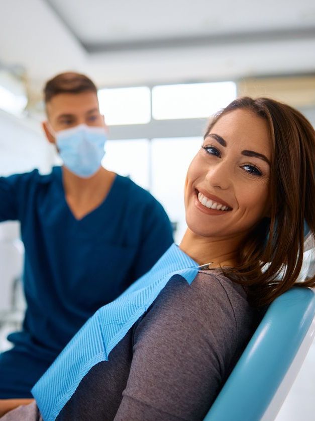 A woman is smiling while sitting in a dental chair next to a dentist wearing a mask.