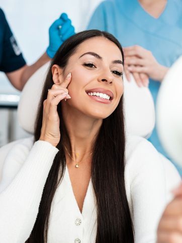 A woman is sitting in a dental chair looking at her face in a mirror.