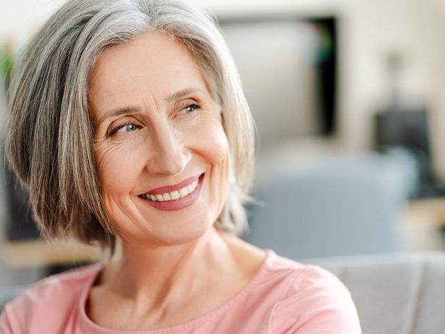 A woman with gray hair is smiling while sitting on a couch.