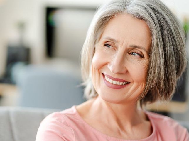 An older woman with gray hair is smiling while sitting on a couch.