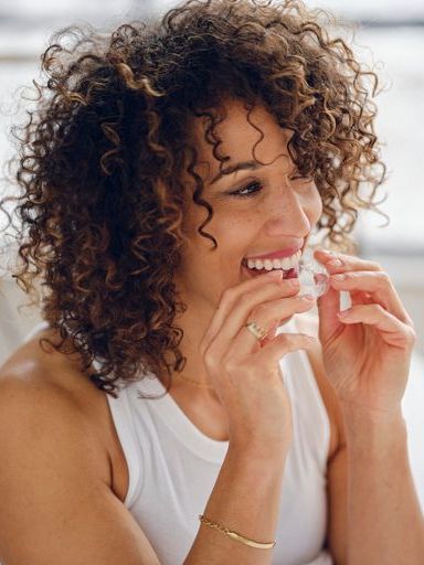 A woman with curly hair is smiling while wearing a white tank top.