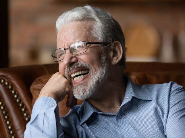 An elderly man with glasses is sitting in a chair and smiling.