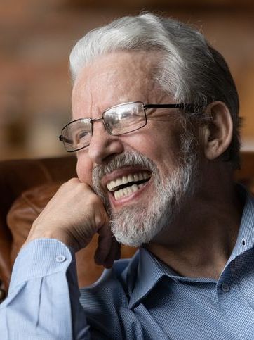 An older man with glasses and a beard is smiling while sitting in a chair.