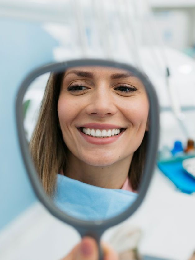 A woman is smiling while looking at her teeth in a hand mirror.