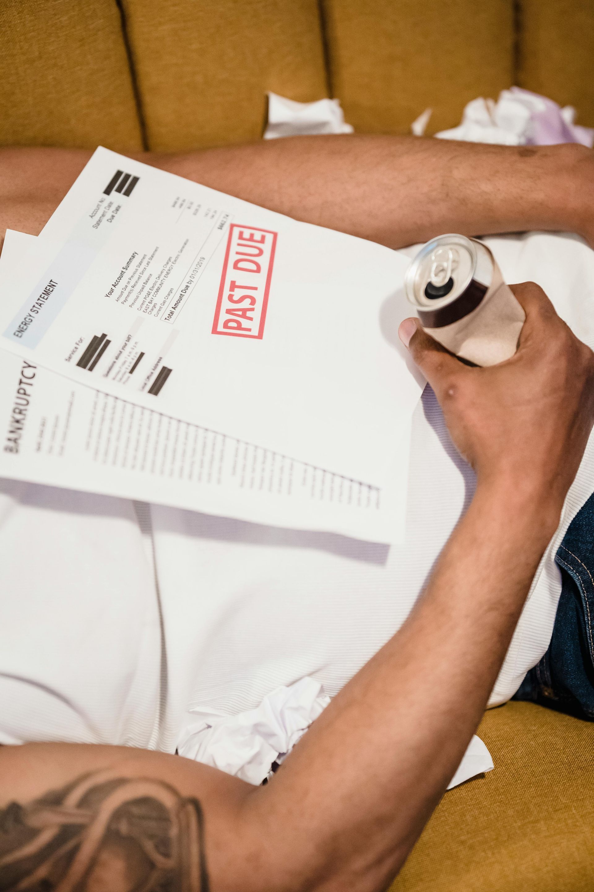 A man is laying on a couch holding a can of beer and a piece of paper that says past due.