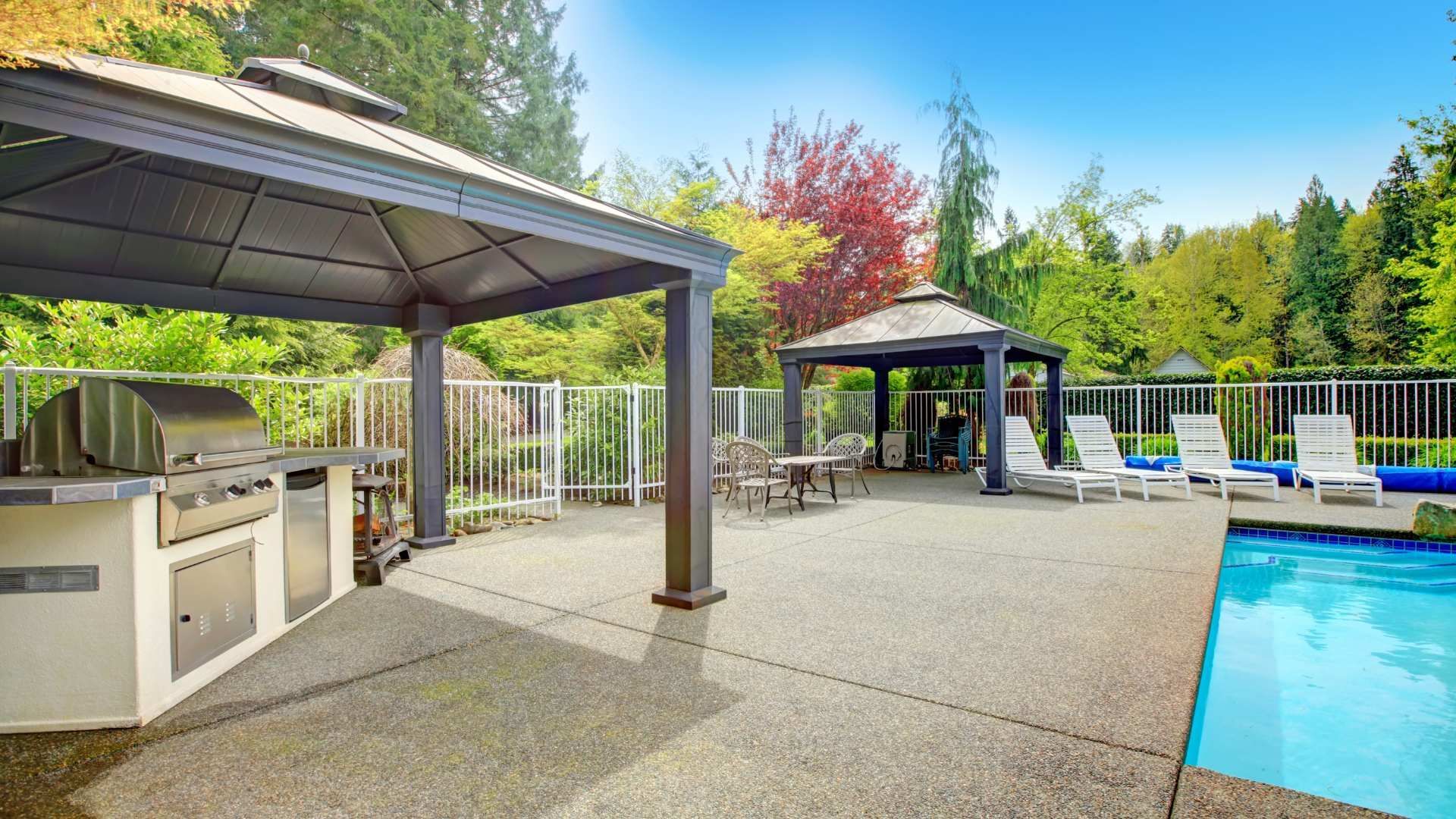 A patio with a gazebo , grill , chairs and a swimming pool.