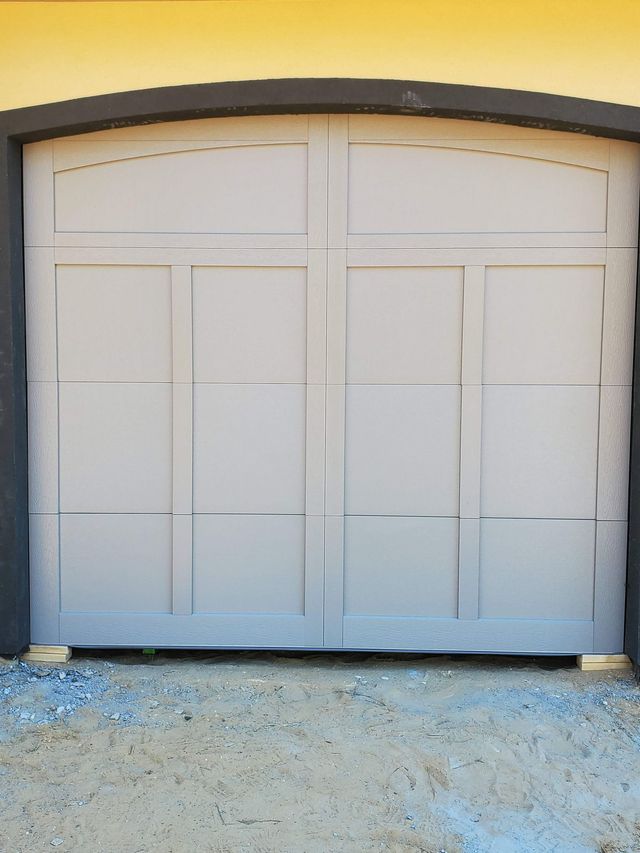 White double garage door with arched top, set in a beige wall above a gravel driveway.