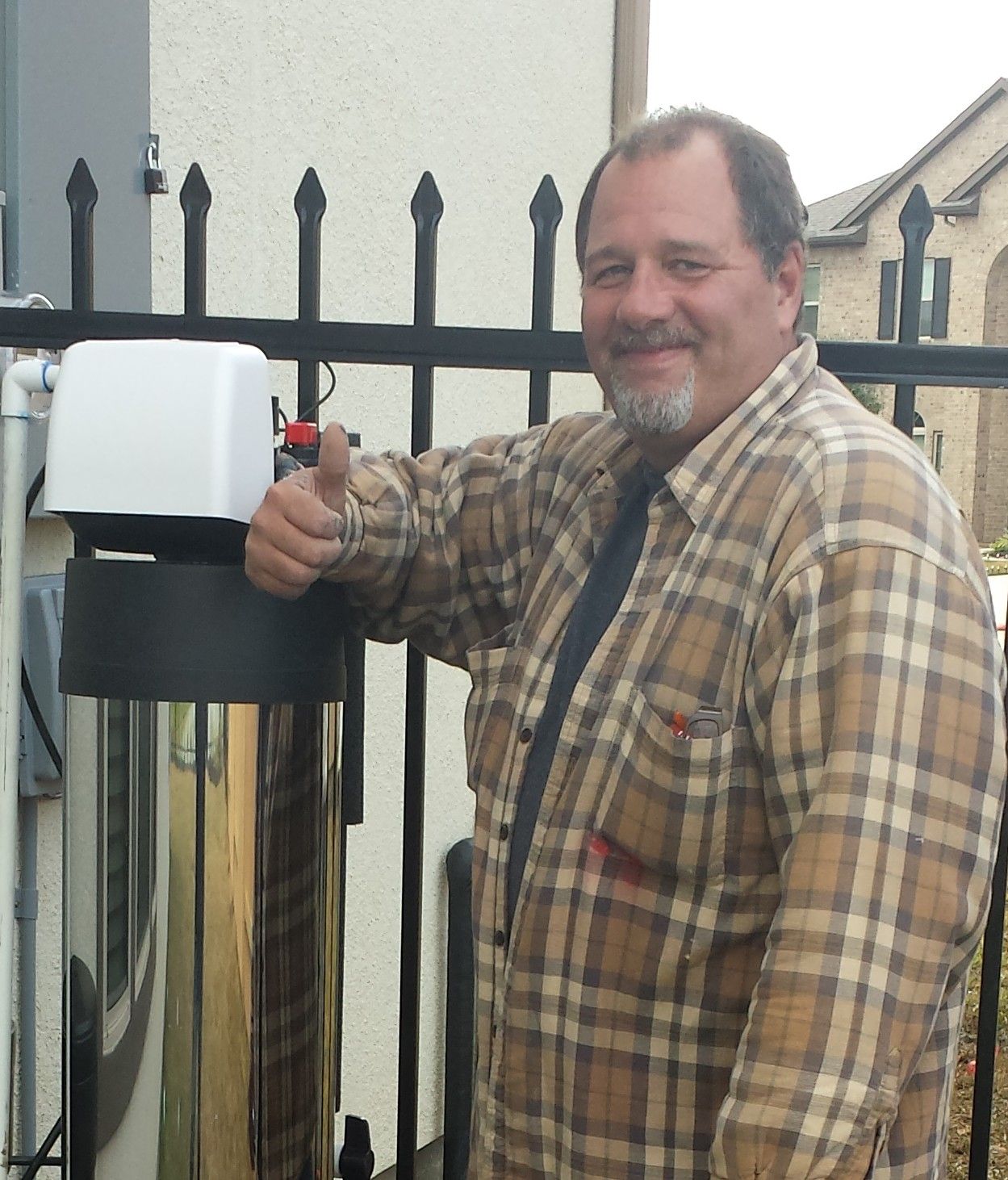 Man giving thumbs up next to a water filtration system outside a house, smiling.