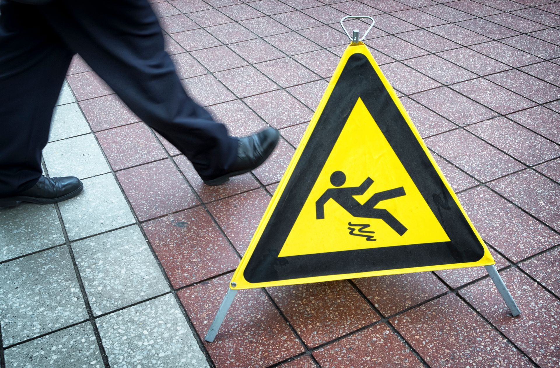 Yellow caution sign with falling person symbol on wet floor. Person walking past.