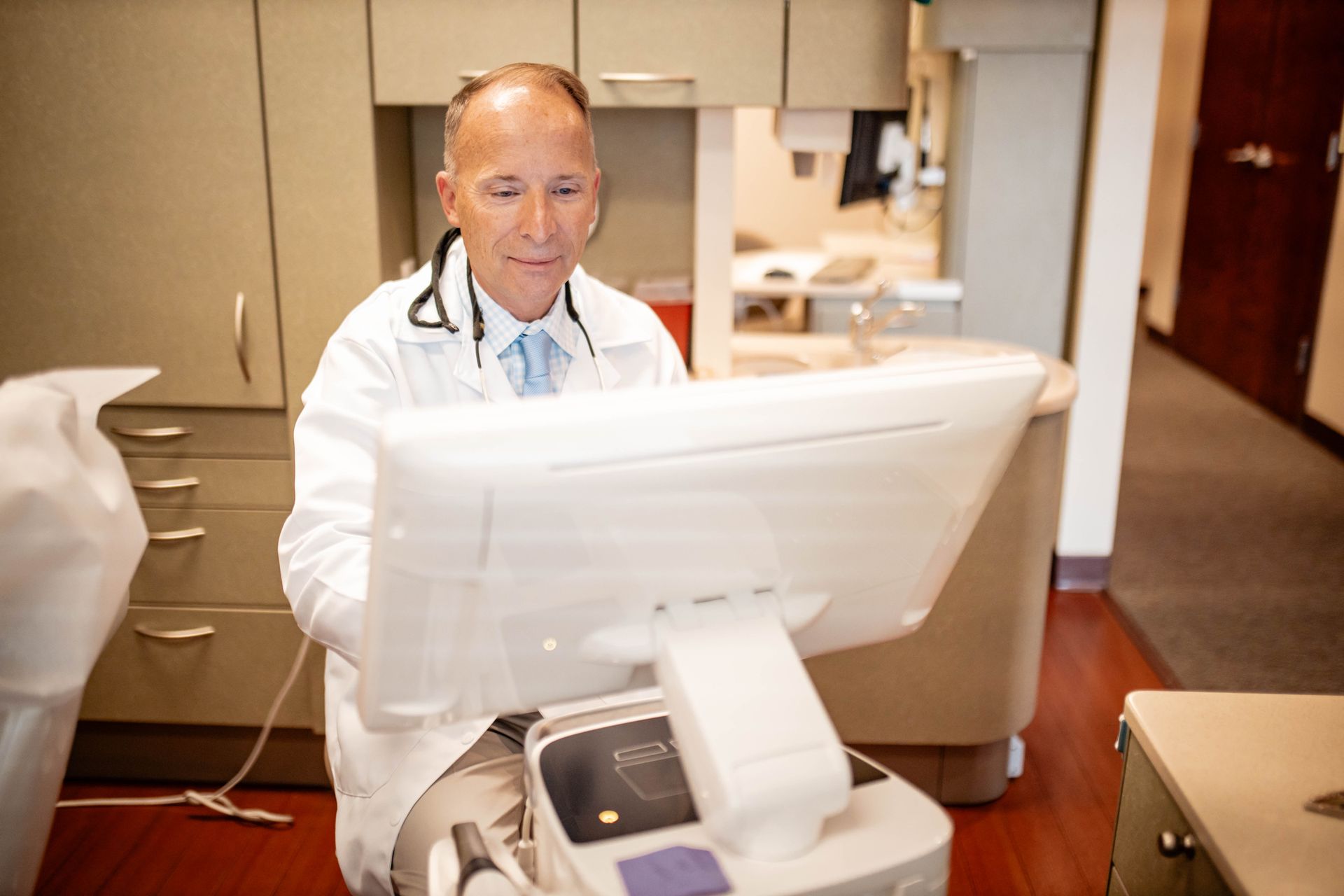 A doctor in a lab coat is sitting in front of a computer monitor.