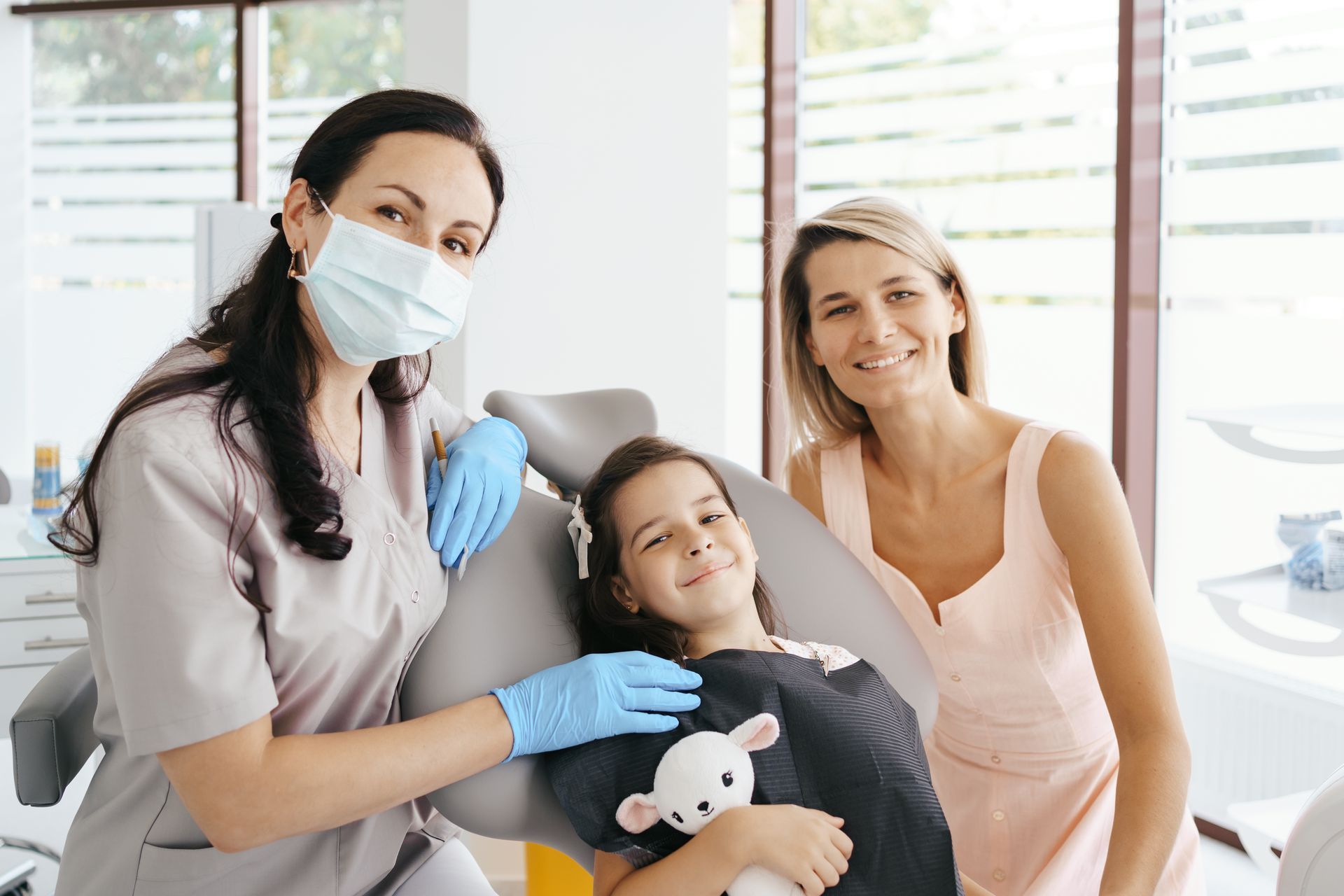 A little girl is sitting in a dental chair with two women.