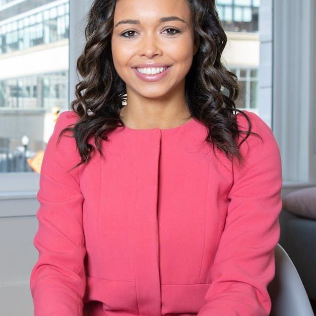 A woman in a pink jacket is smiling while sitting in front of a window.
