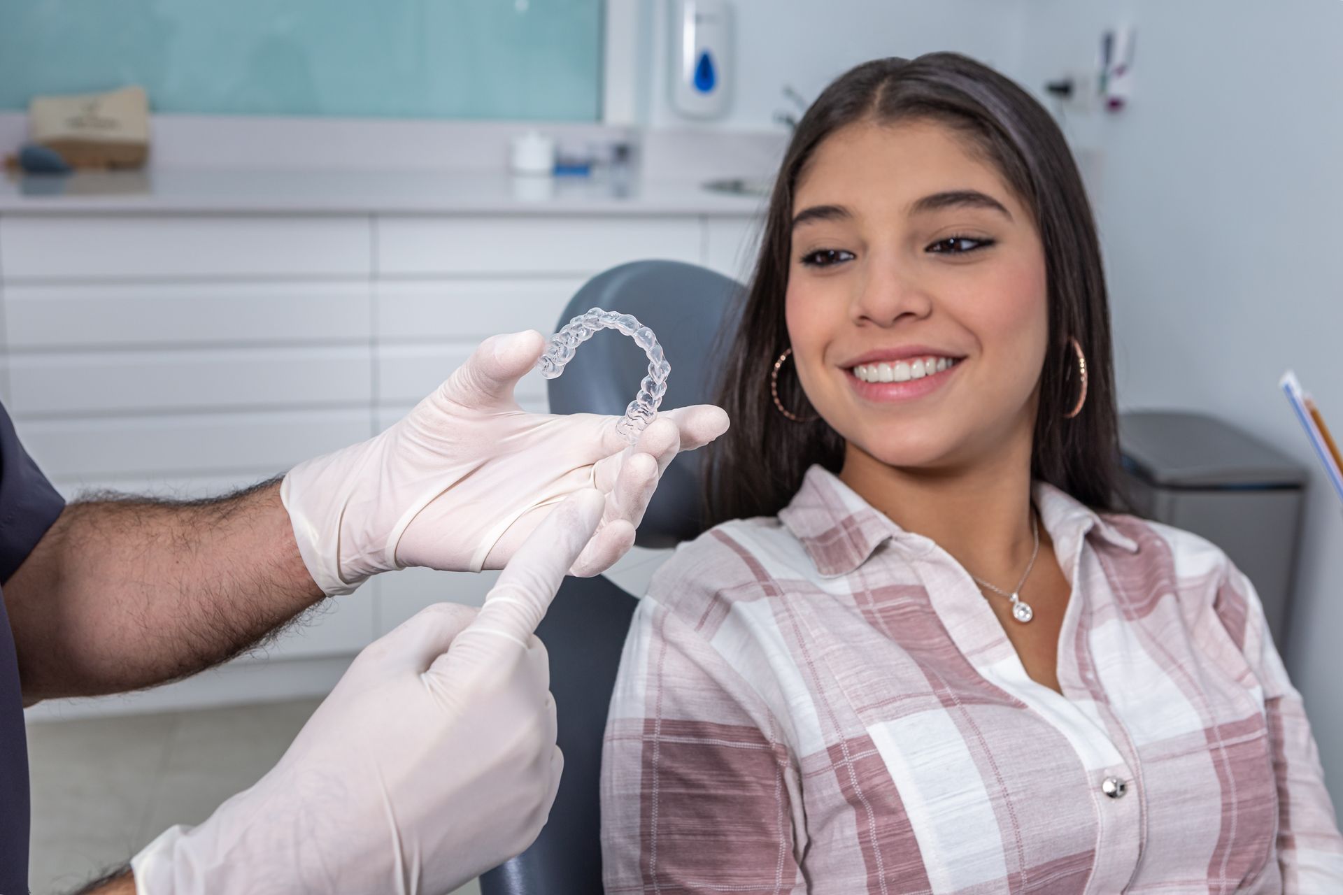 A woman is sitting in a dental chair while a dentist holds a clear retainer in his hand.