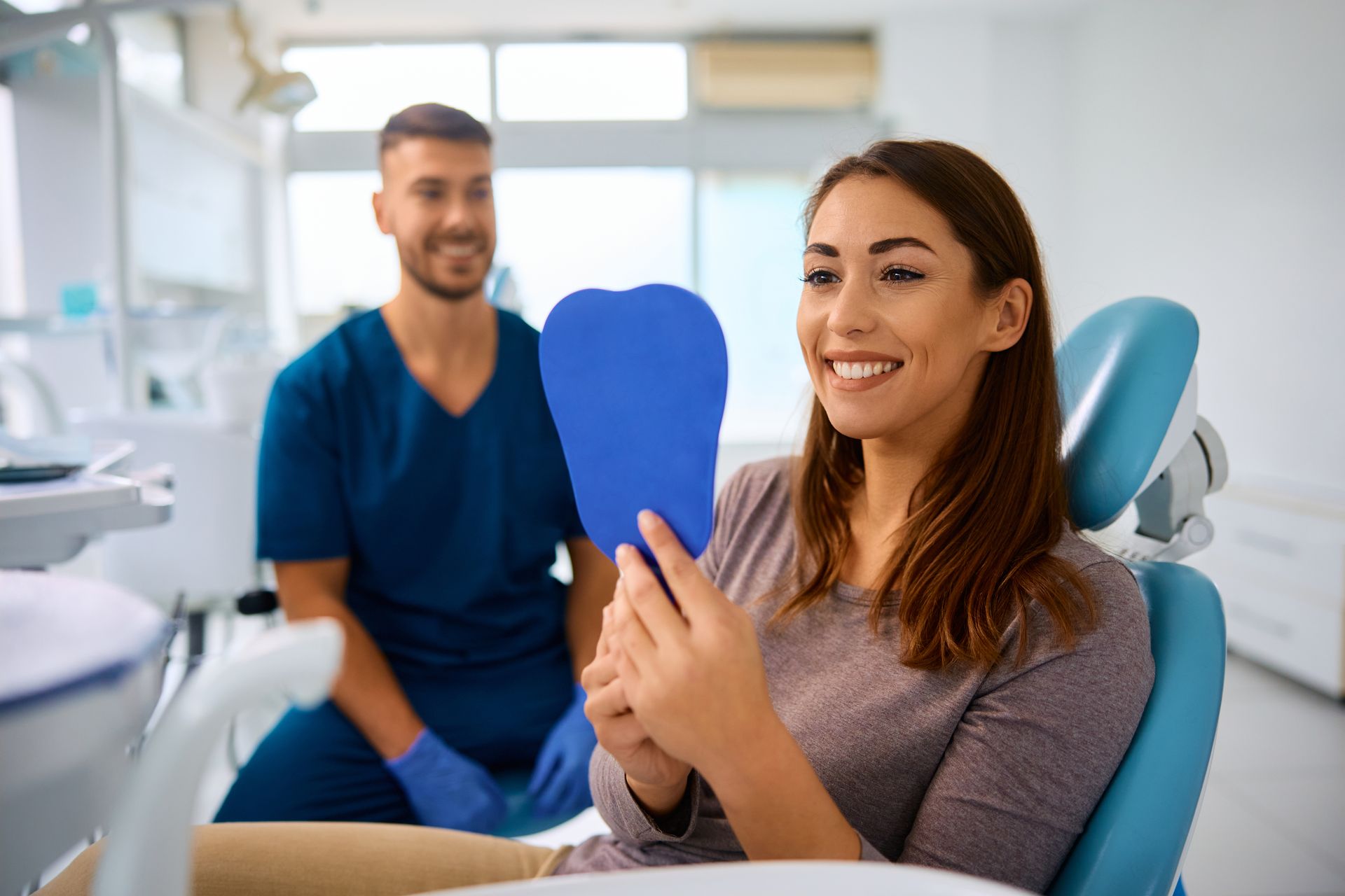 A woman is sitting in a dental chair looking at her teeth in a mirror.