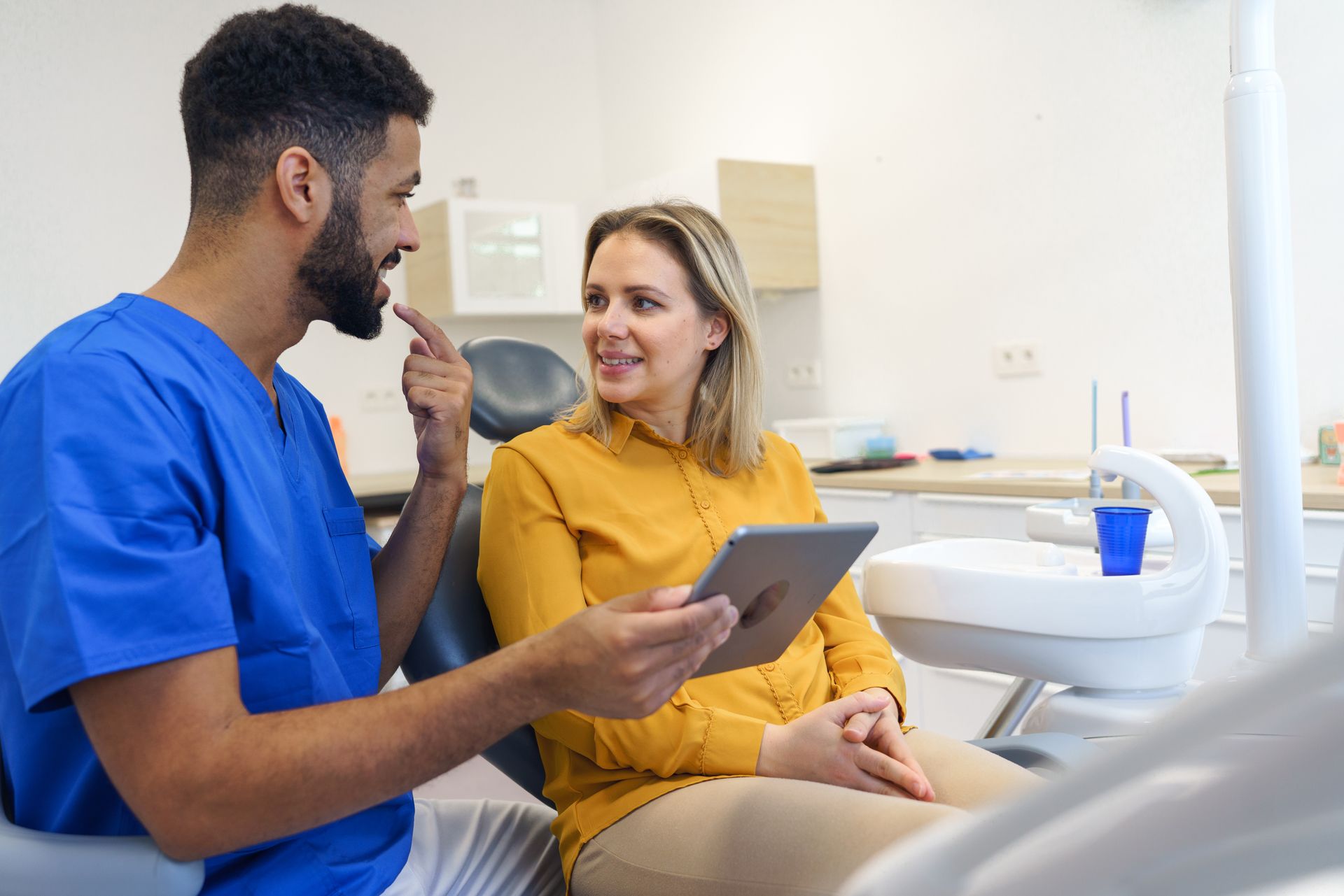 A dentist is talking to a patient in a dental chair while holding a tablet.