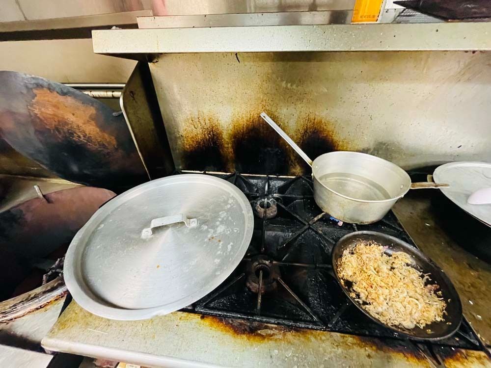 A dirty commercial kitchen stovetop with a pan of food, a pot of water, and a lid.