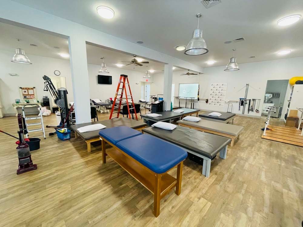 Physical therapy room with multiple treatment tables, various exercise equipment, and overhead lighting.