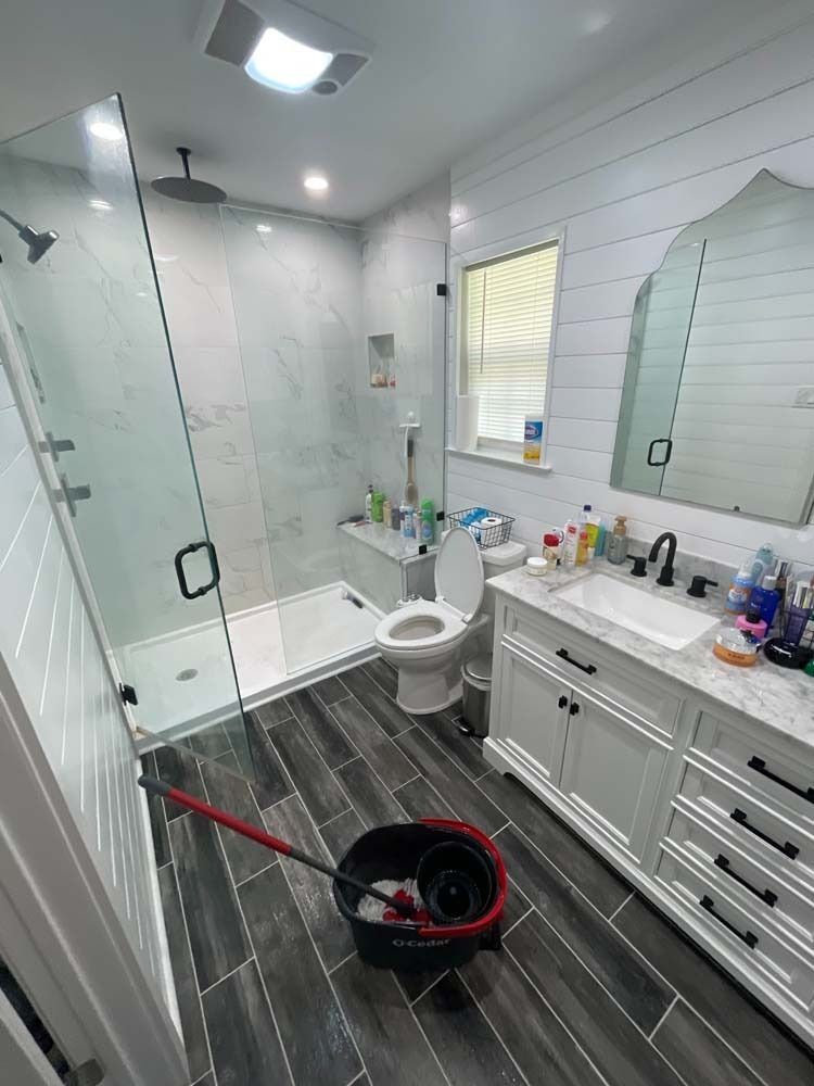 Bathroom being cleaned, with mop and bucket in foreground. Shower, toilet, and vanity are visible.