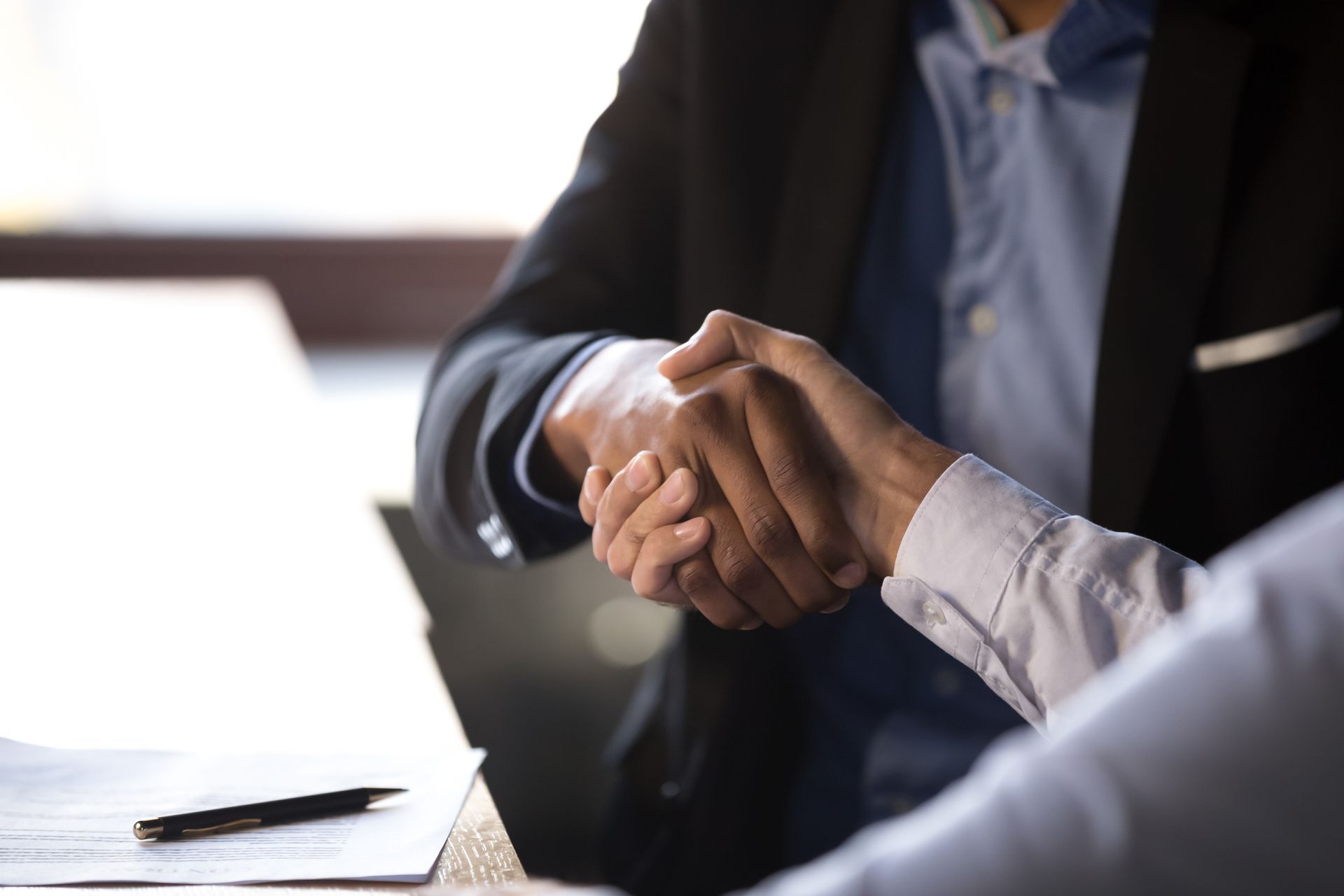 Two people in business attire shaking hands over a table with a pen and papers.