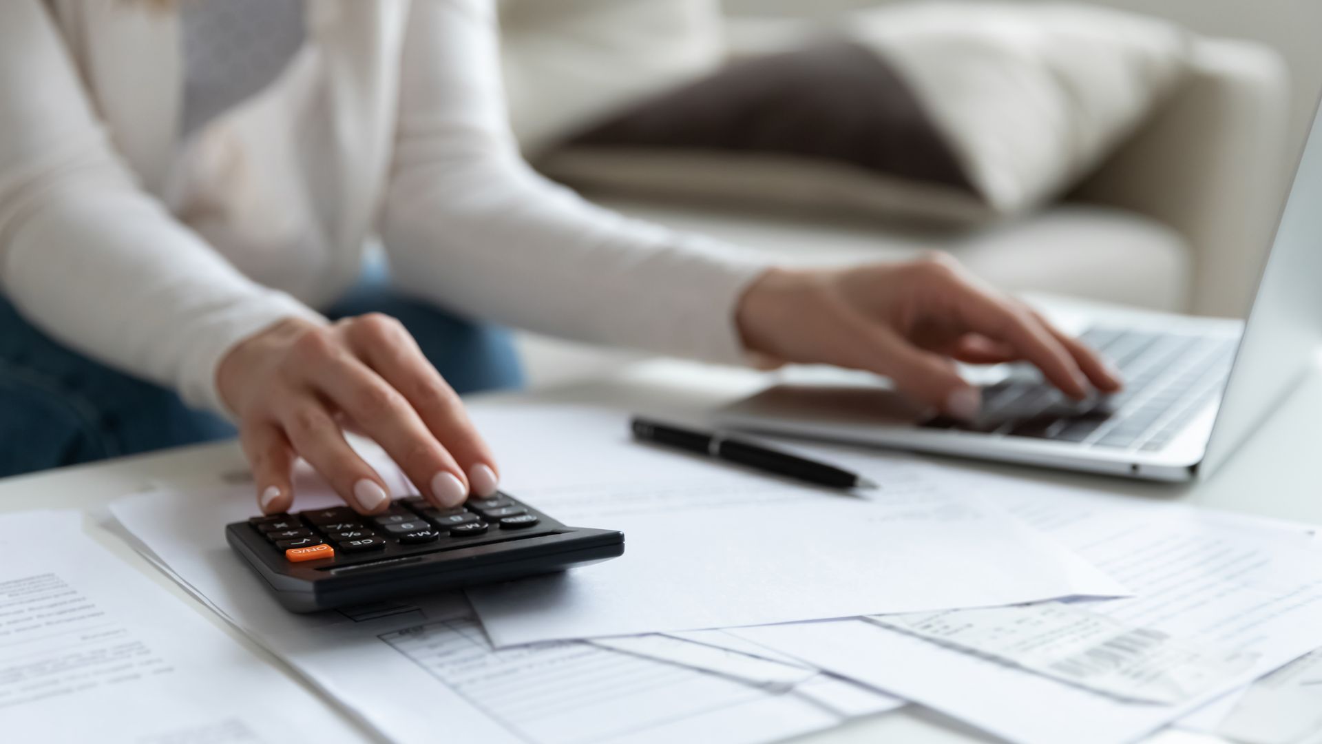 Woman using a calculator and laptop, reviewing documents on a table.