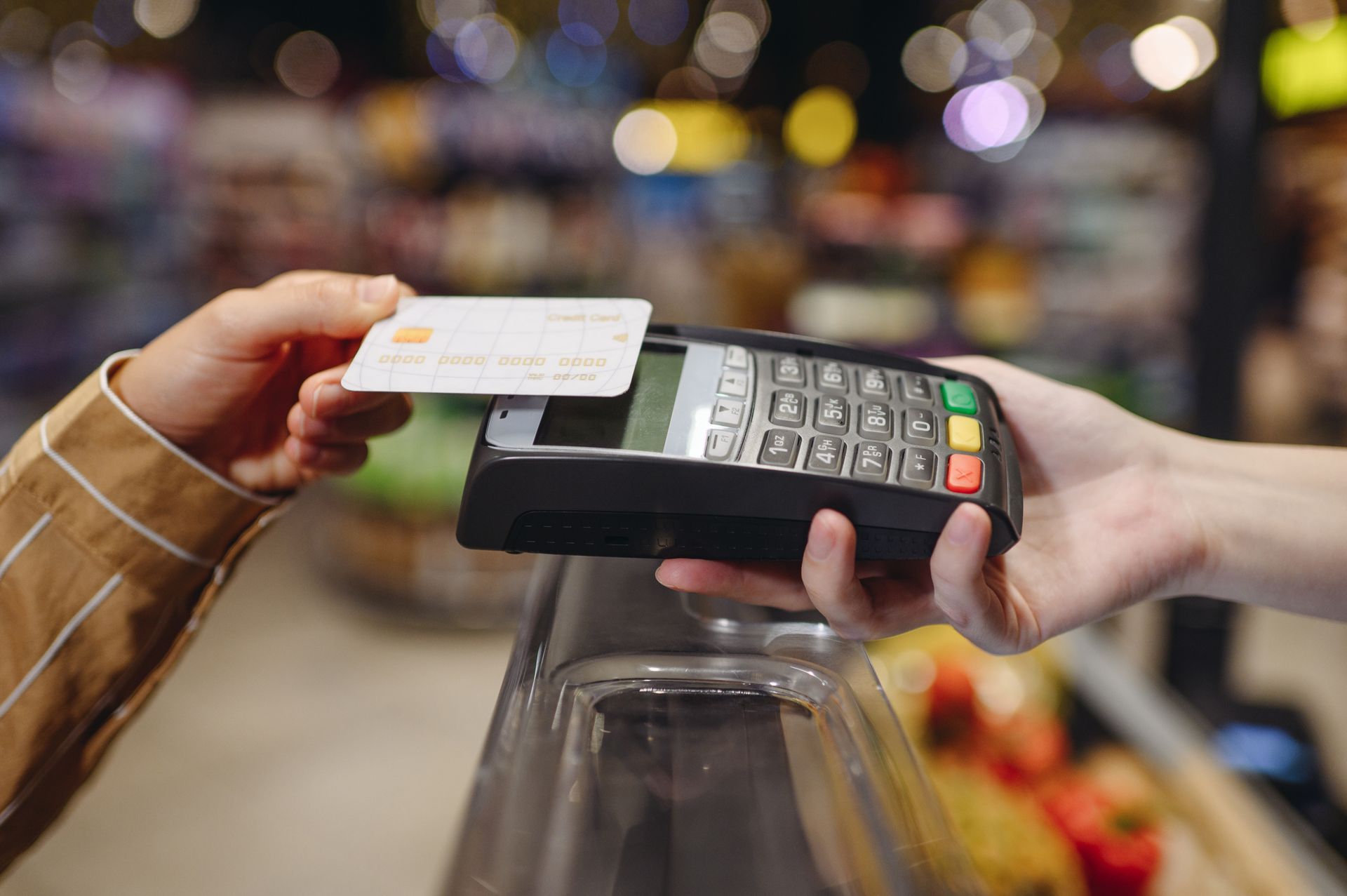 Hand holding a white credit card over a card reader in a grocery store.