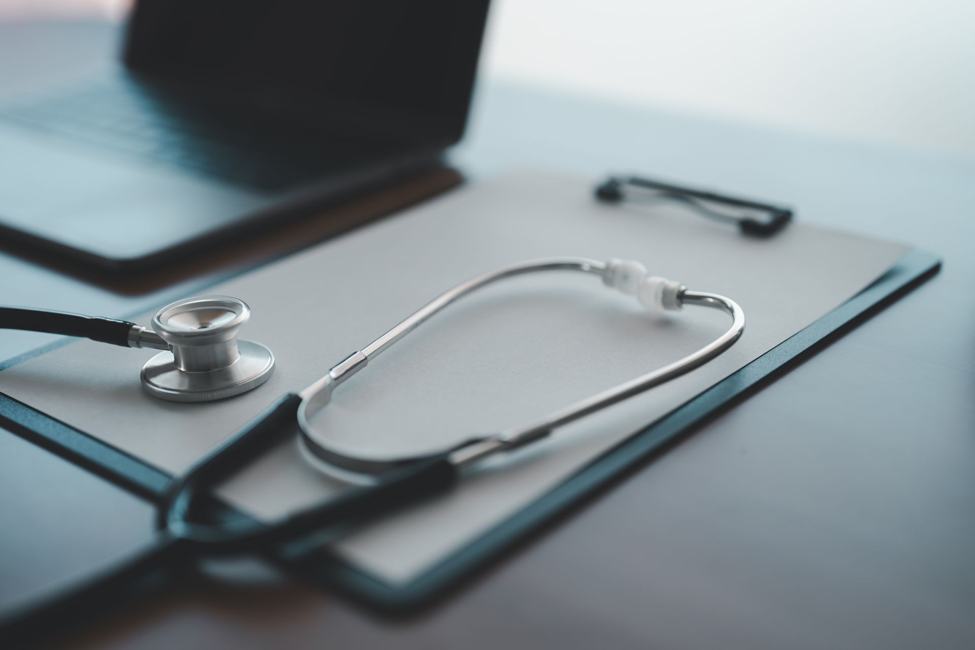 Stethoscope on clipboard next to a laptop, likely in a medical setting.