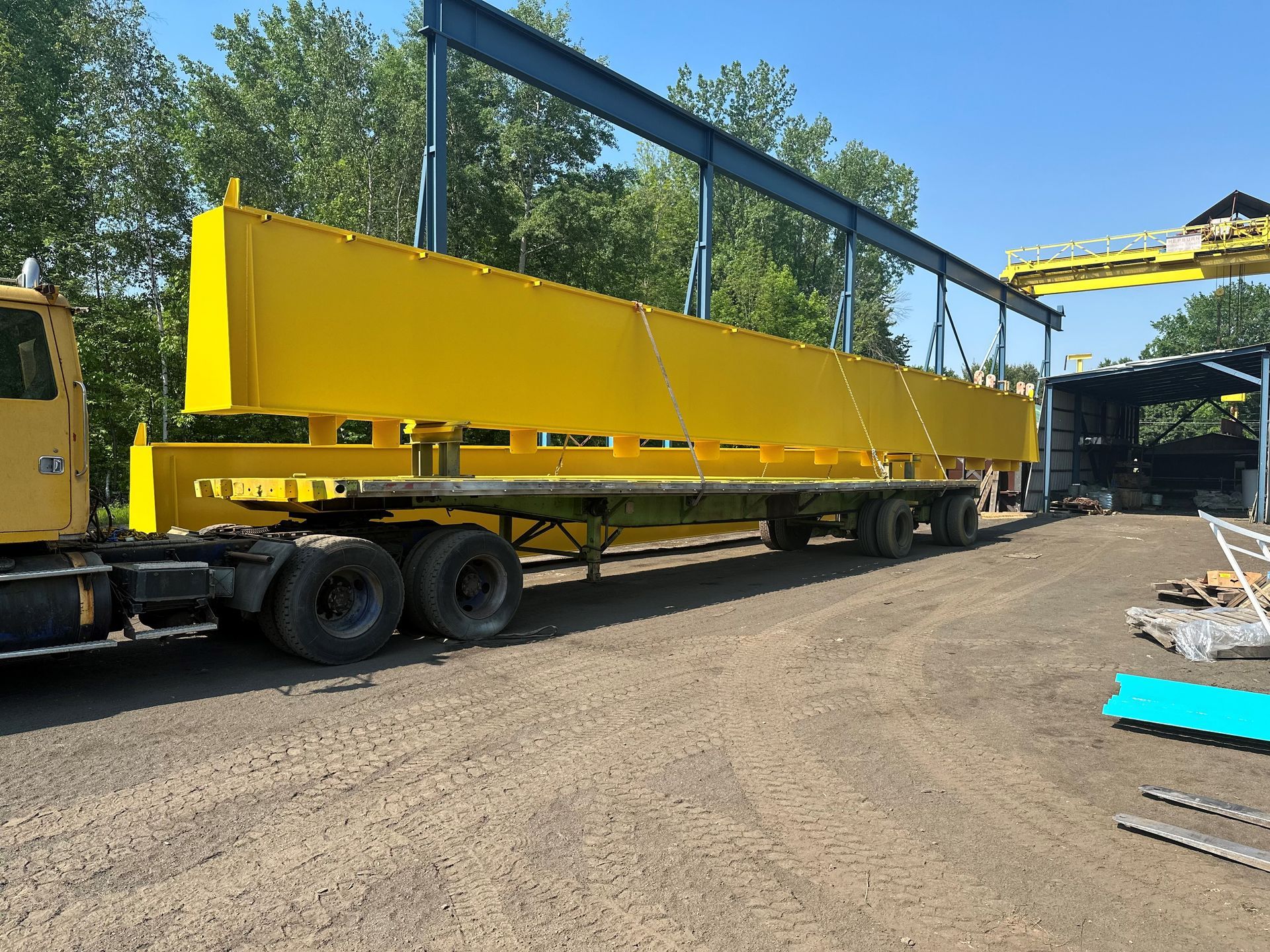 Poutre jaune chargée sur une remorque, suspendue par une grue dans un environnement industriel.