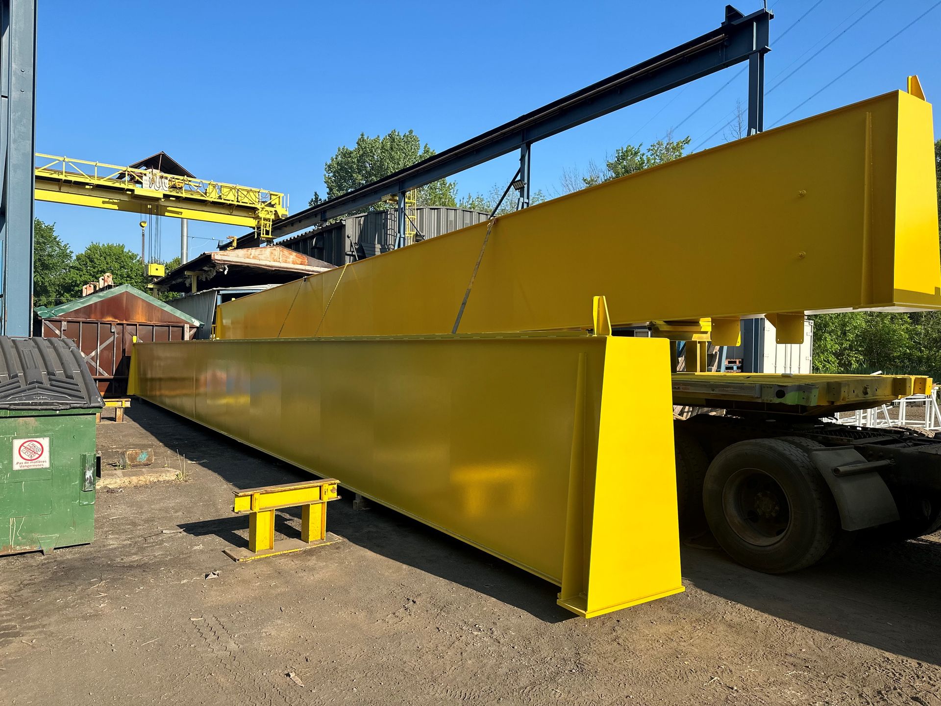 Poutres de pont roulant jaunes sur une remorque à l'extérieur d'un bâtiment, sous un ciel bleu.