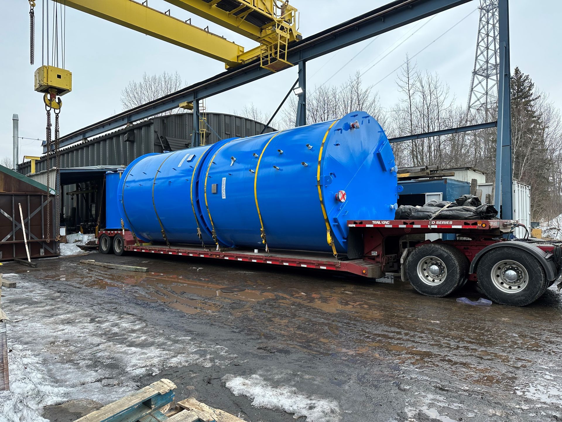 Citerne industrielle bleue sur un camion à plateau, déchargée sous une grue jaune dans un environnement industriel enneigé.