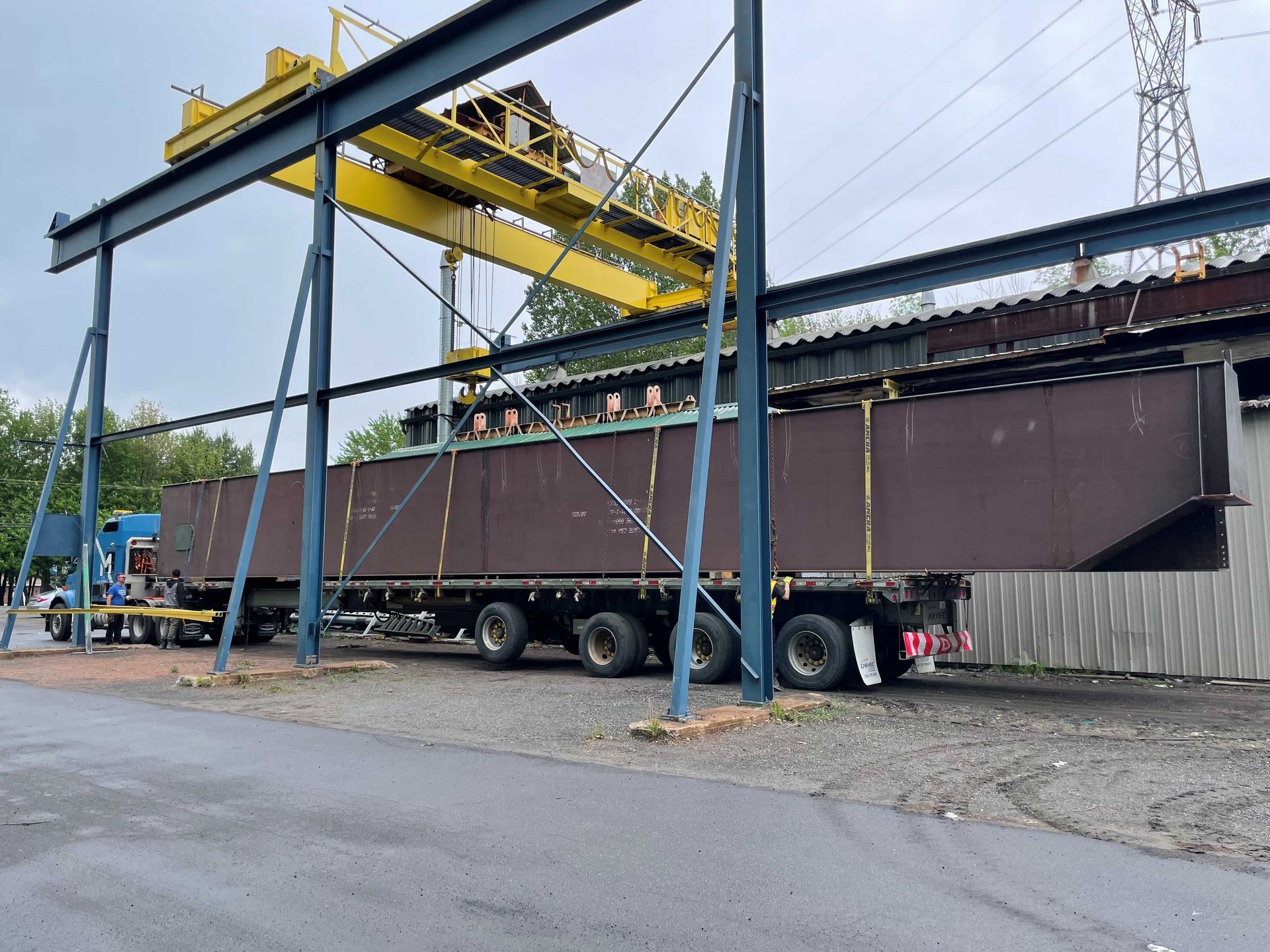 Une grande poutre en acier marron est chargée sur un camion sous une grue jaune sur un site industriel.