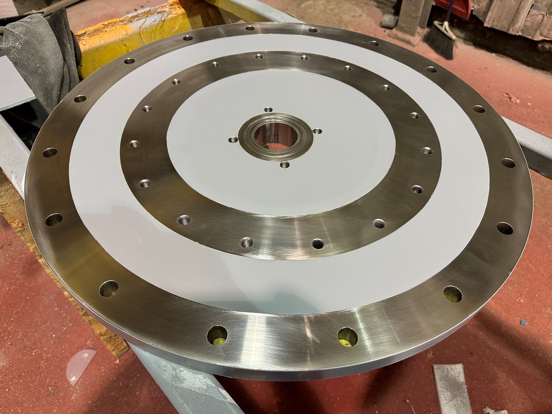 Composant métallique circulaire avec anneaux concentriques blancs et argentés. Bords boulonnés et moyeu central.