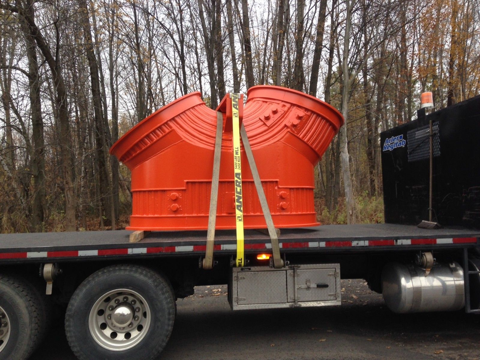 Structure métallique orange fixée sur un camion à plateau avec des sangles jaunes, arbres en arrière-plan.