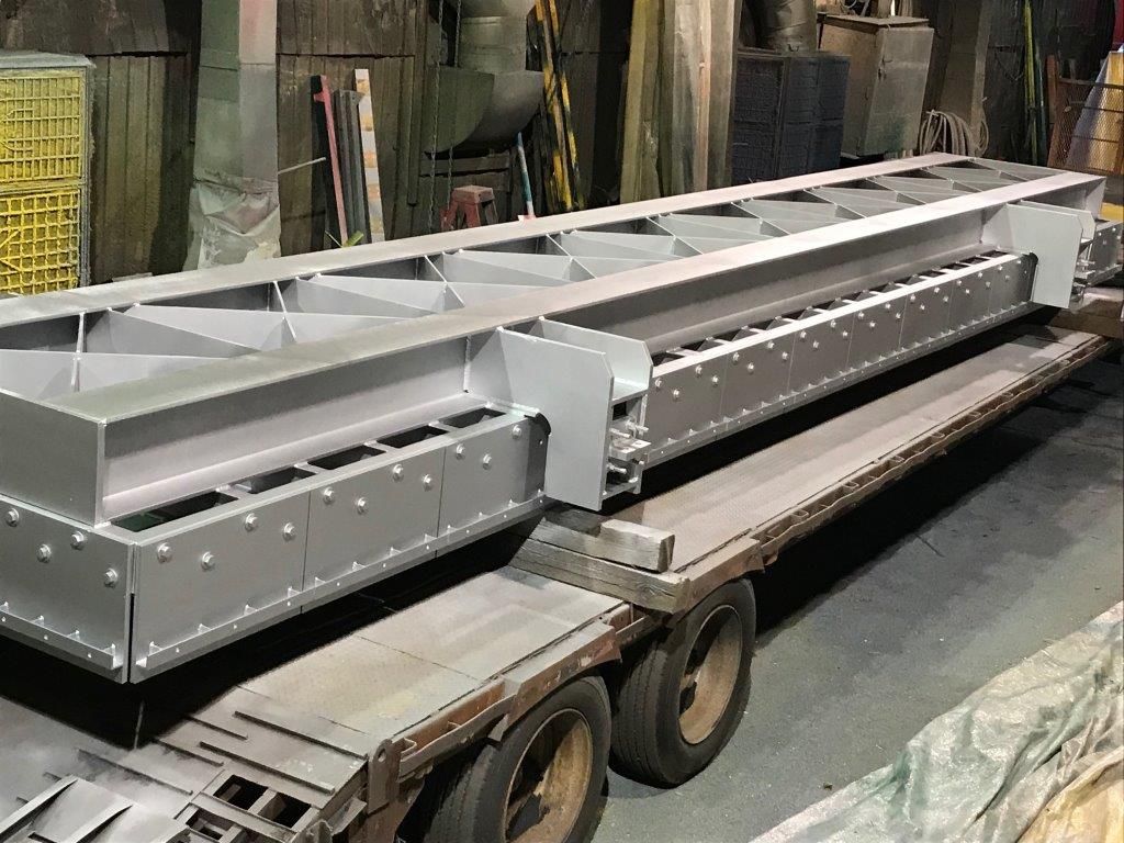 Une grande structure métallique grise sur une remorque, peut-être un composant de convoyeur, à l'intérieur d'une usine.