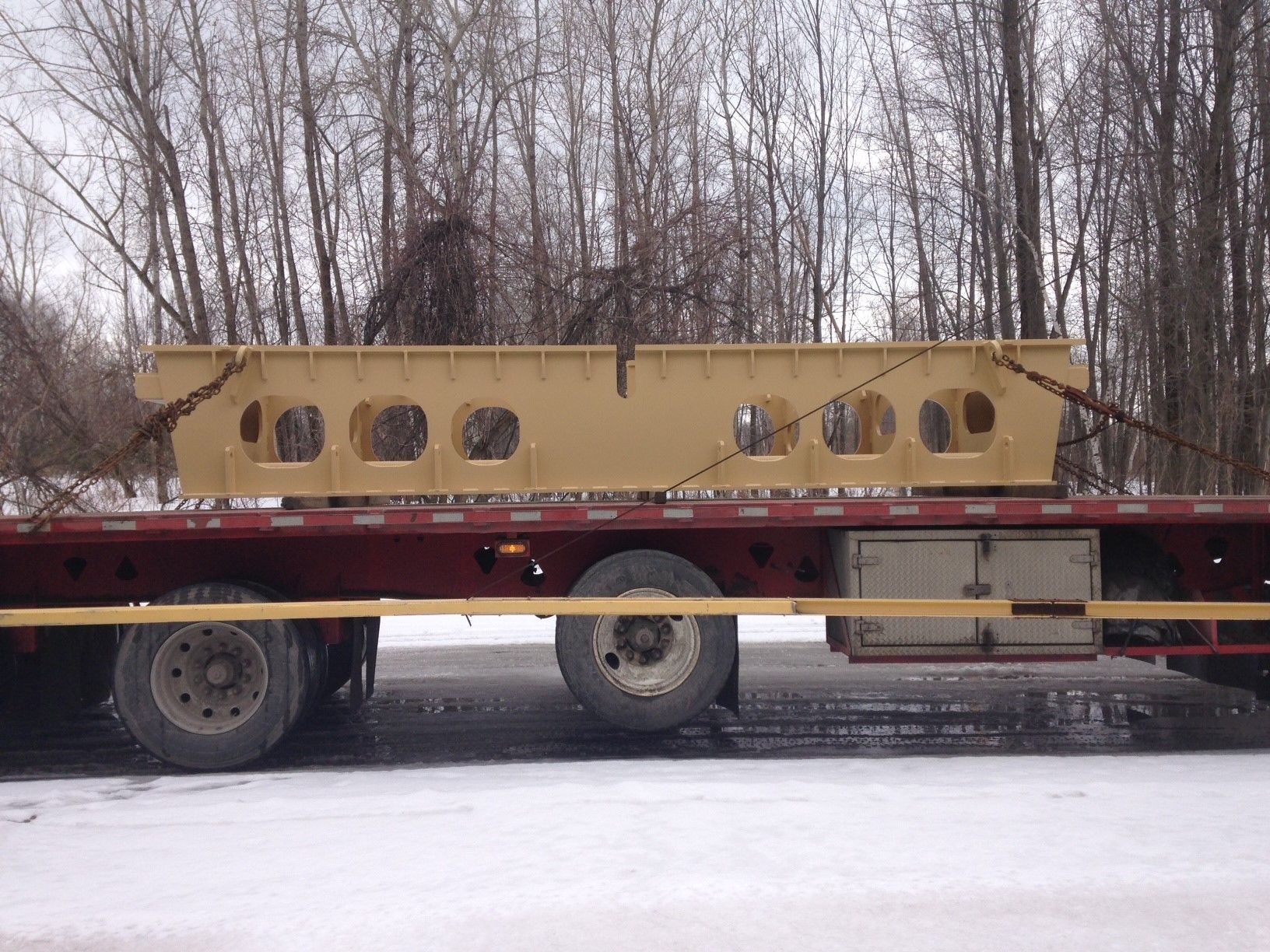Une grande poutre métallique beige fixée à une remorque à plateau sur une route enneigée, avec des arbres en arrière-plan.