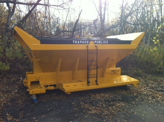 Épandeur de sel jaune pour travaux publics, texte noir, échelle et décor extérieur.