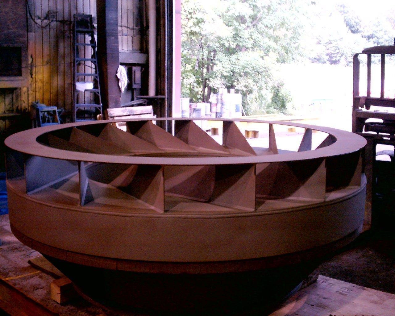 Grande roue de turbine, en métal gris avec pales inclinées, à l'intérieur près d'une fenêtre.