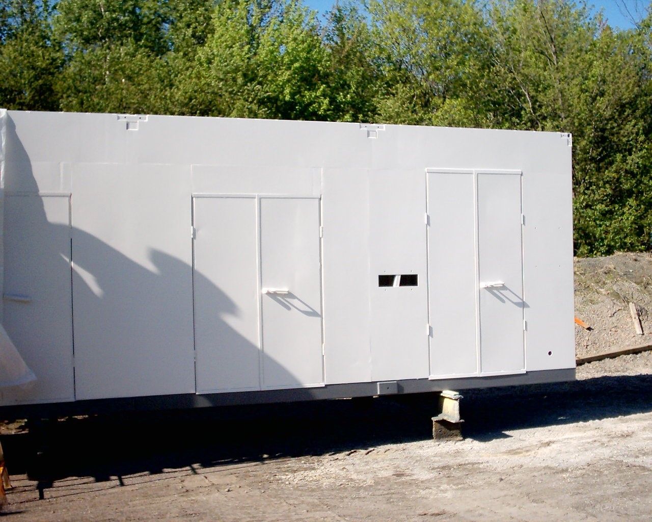 Bâtiment rectangulaire blanc avec deux portes, de petites fenêtres et des arbres verts en arrière-plan.