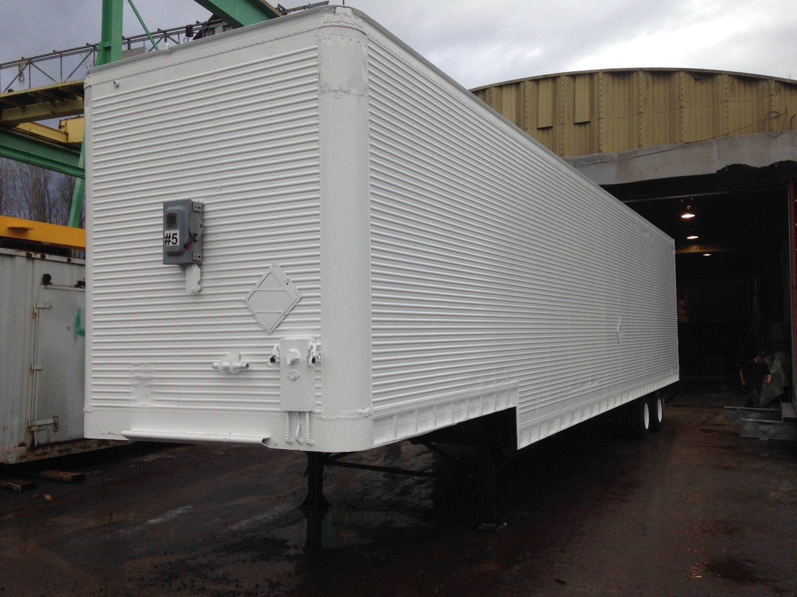 Semi-remorque blanche avec revêtement en tôle ondulée, stationnée sur une surface mouillée, partiellement à l'intérieur d'un bâtiment.