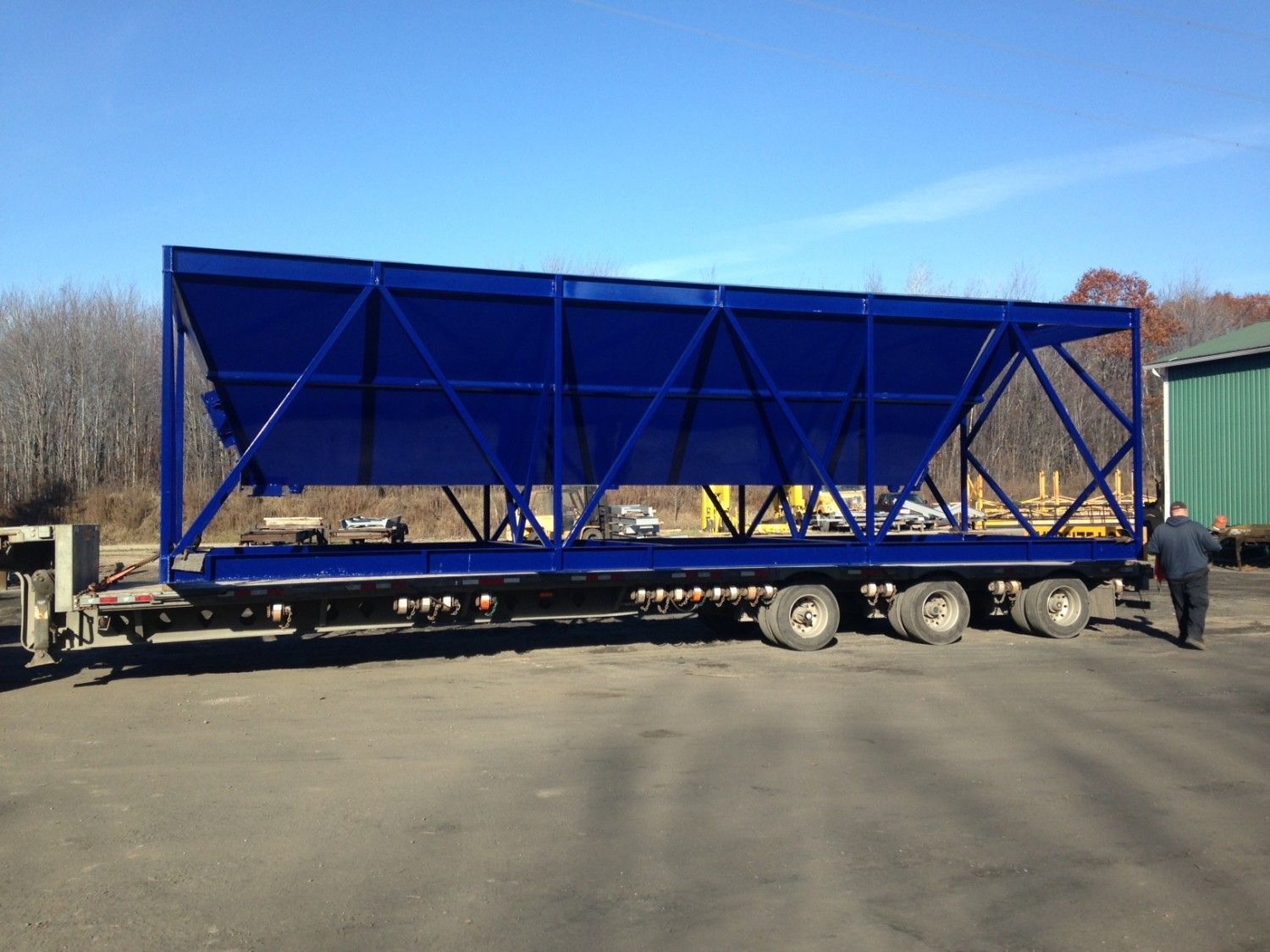 Trémie industrielle bleue sur une remorque, en extérieur. Un homme se tient à côté, sous un ciel bleu.