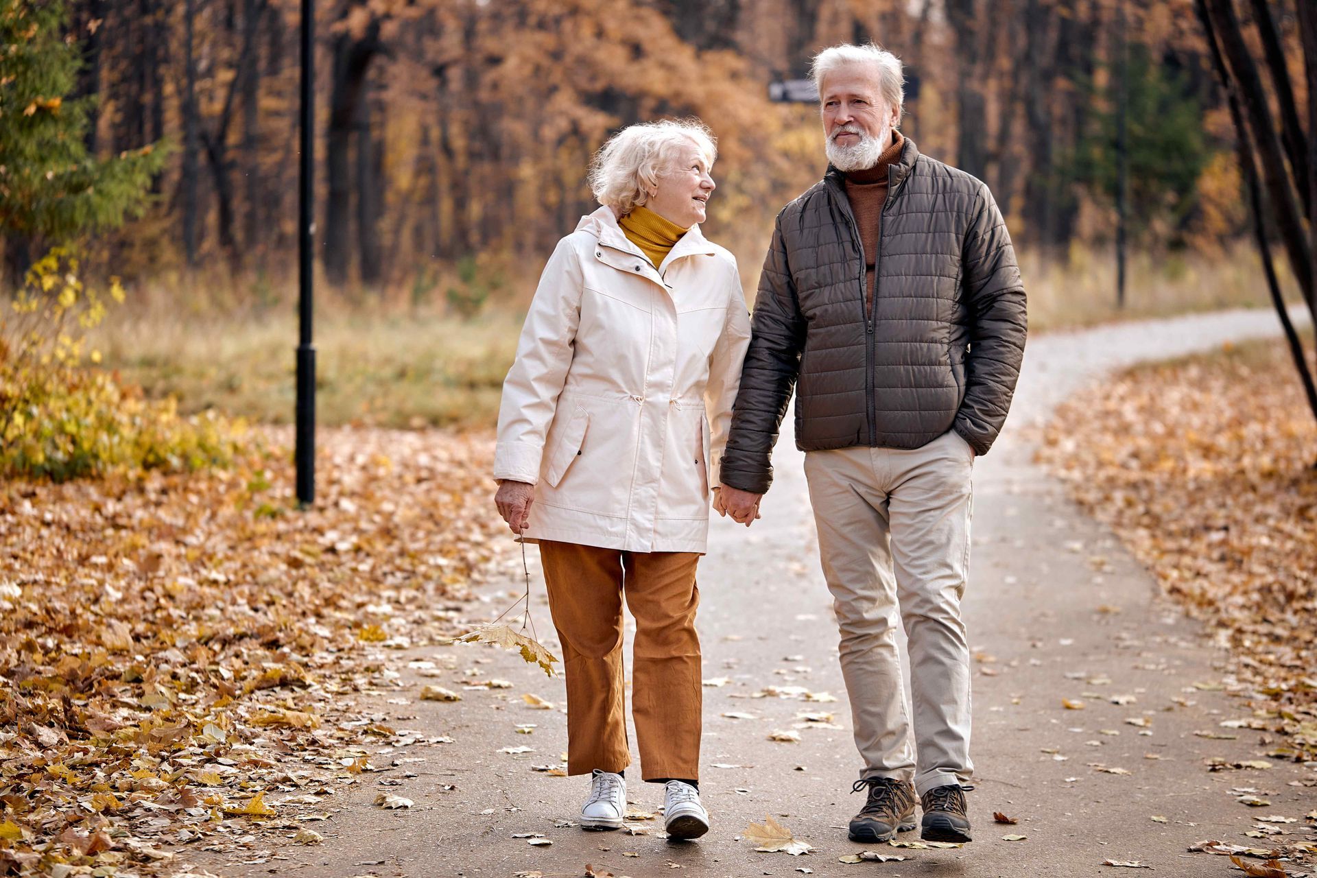 An elderly couple is walking down a path in a park holding hands.
