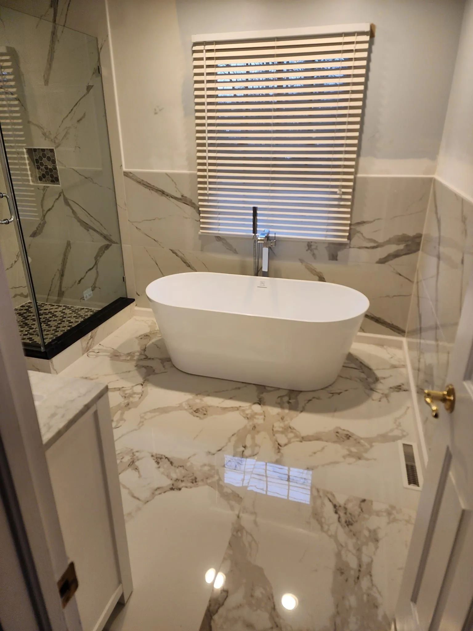 Luxurious bathroom with white, marble-look tile, a freestanding tub, and a window with blinds.