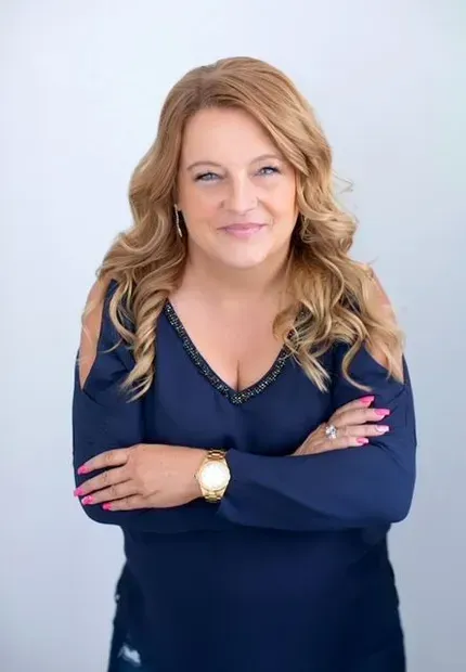 Woman with blonde hair in a blue dress smiles, arms crossed.