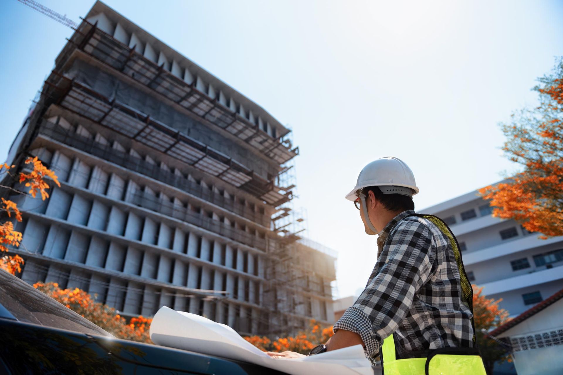 Construction worker in white helmet examines blueprints at a building site under a sunny sky.