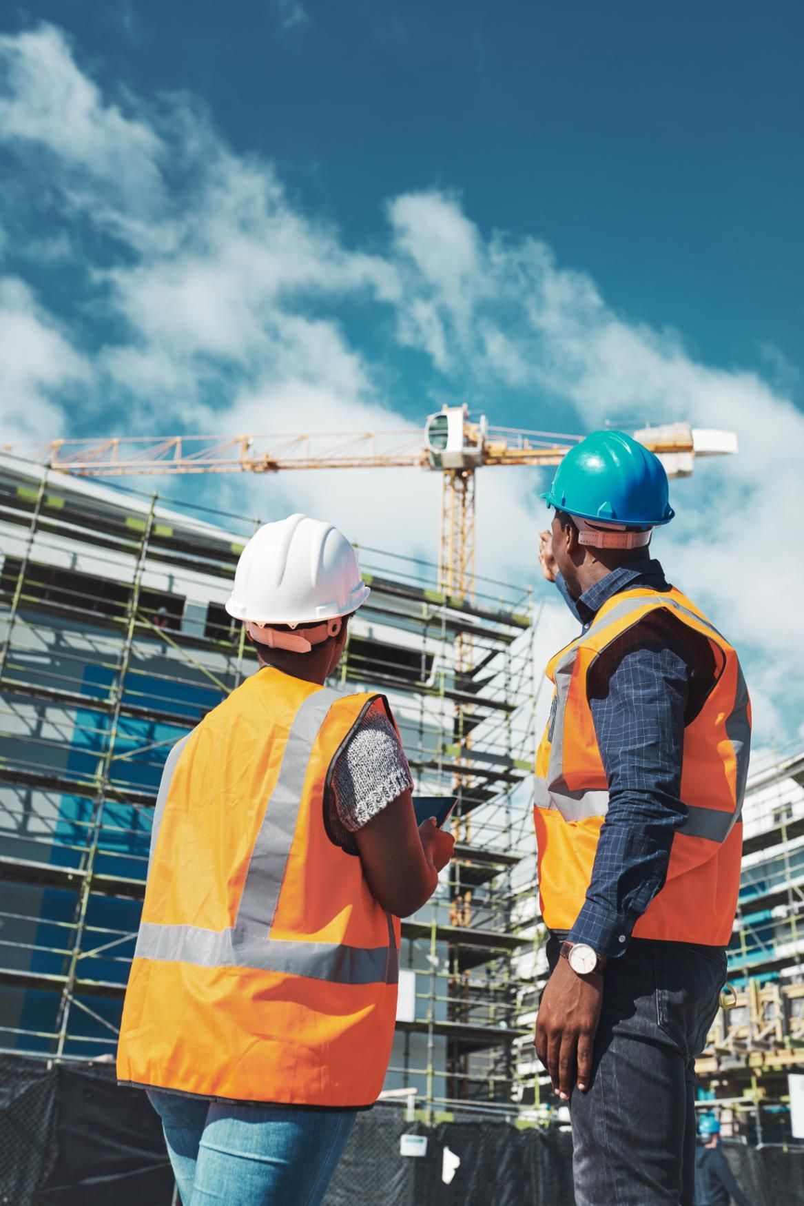 Two construction workers, wearing hard hats and vests, discuss plans at a building site with a crane.