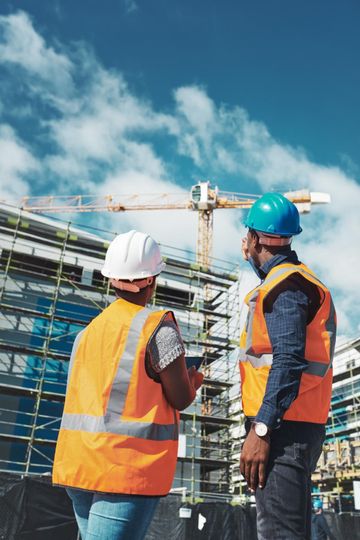 Two construction workers, wearing hard hats and vests, discuss plans at a building site with a crane.