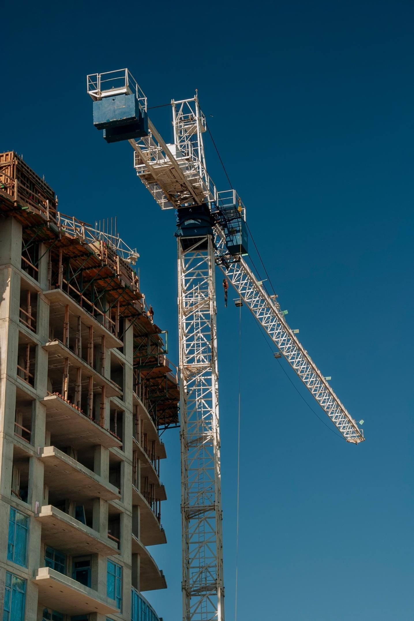 Construction site with a tall crane next to a partially built high-rise against a clear blue sky.