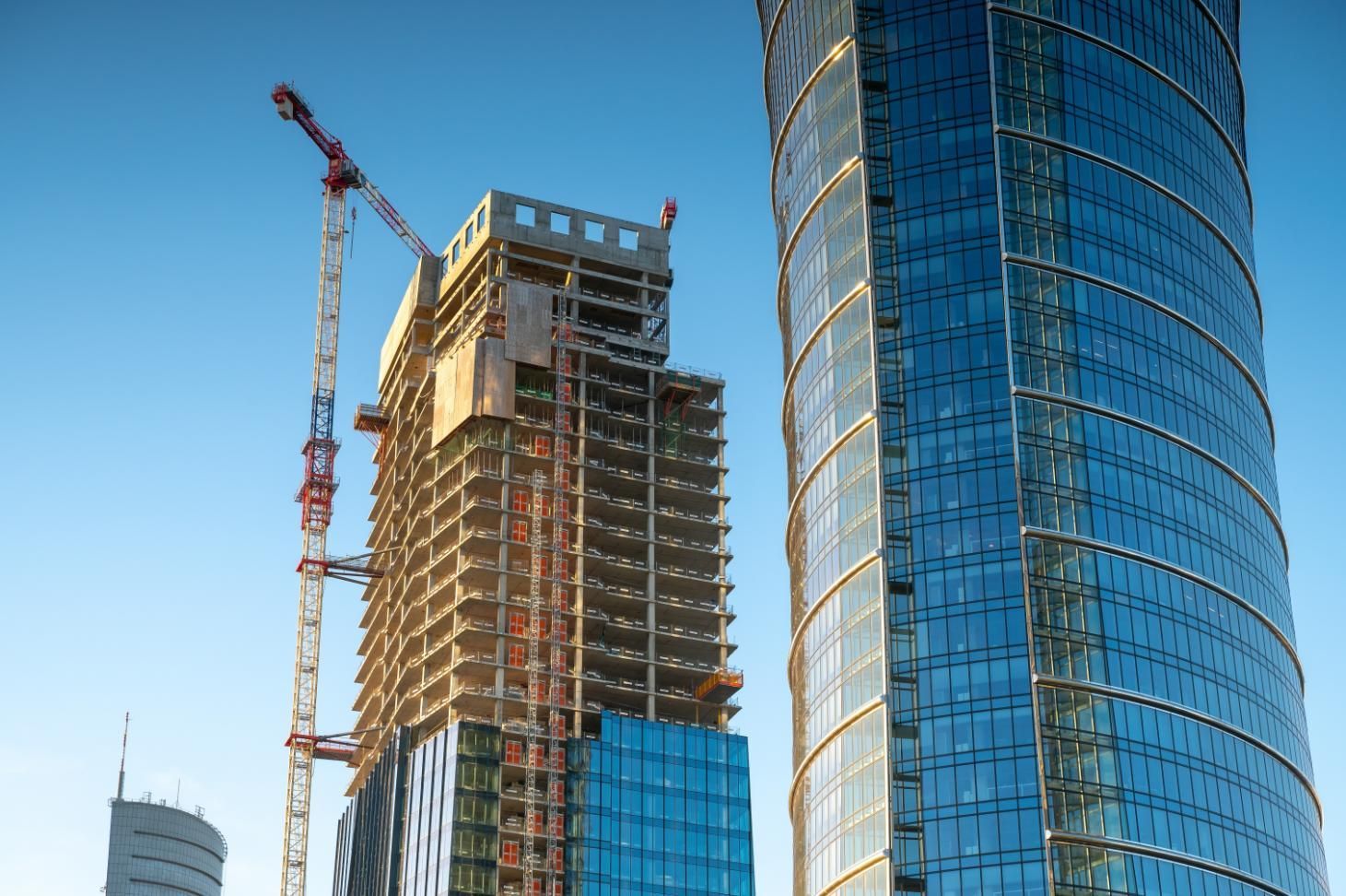 Skyscrapers under construction with cranes against a blue sky. One building is completed, reflecting the sky.